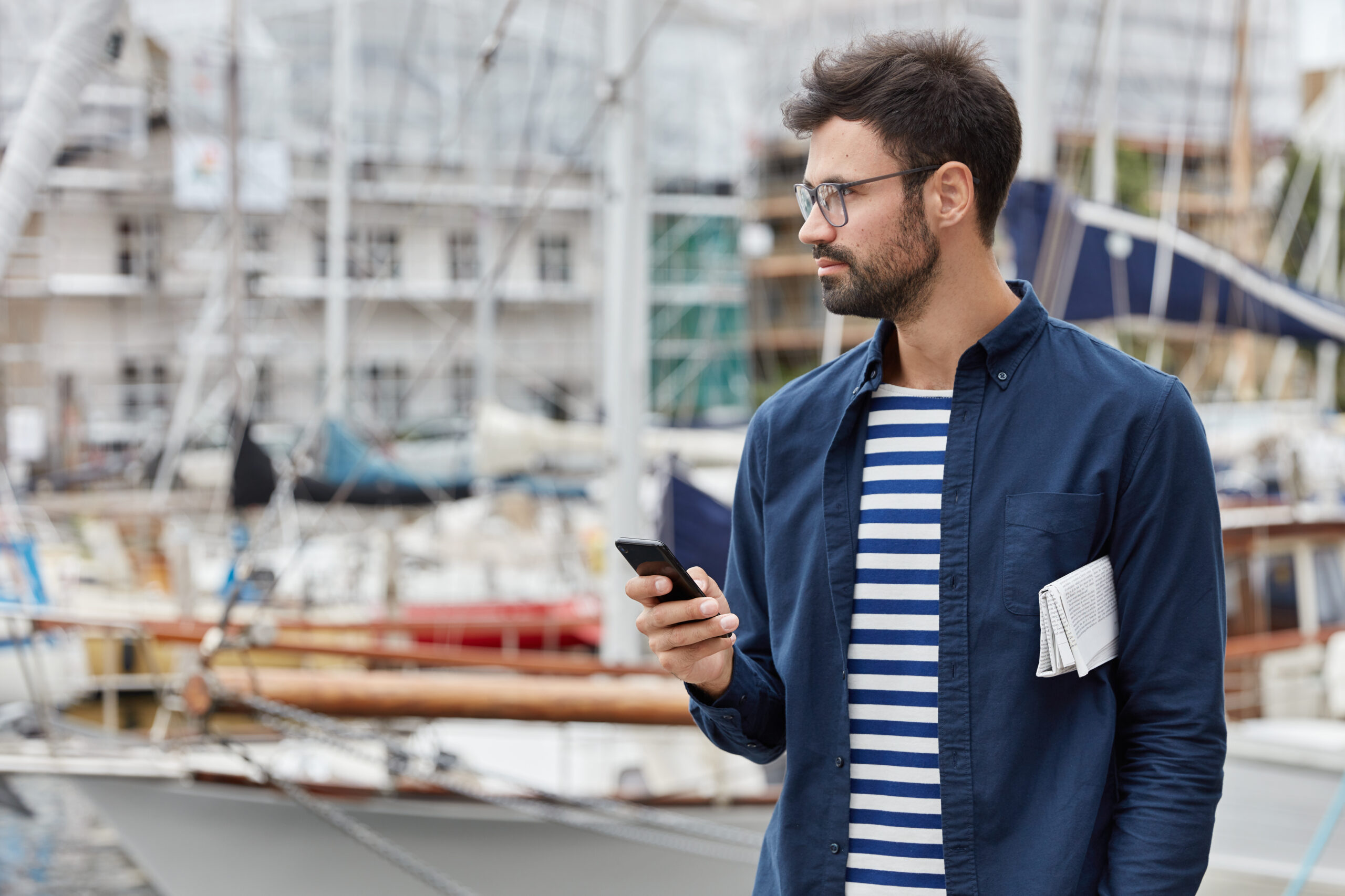 Horizontal view of thoughtful freelancer blogs in social networks, holds contemporary mobile phone, wears transparent glasses, poses against harbour background with copy space for your promotion